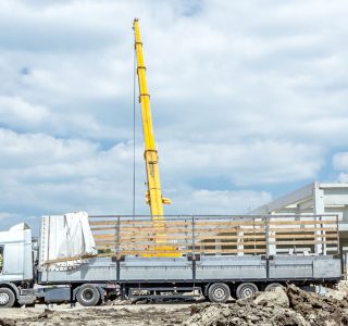 Mobiele kraan tilt een stalen constructiebalk uit een vrachtwagen bij de bouw van een bedrijfshal