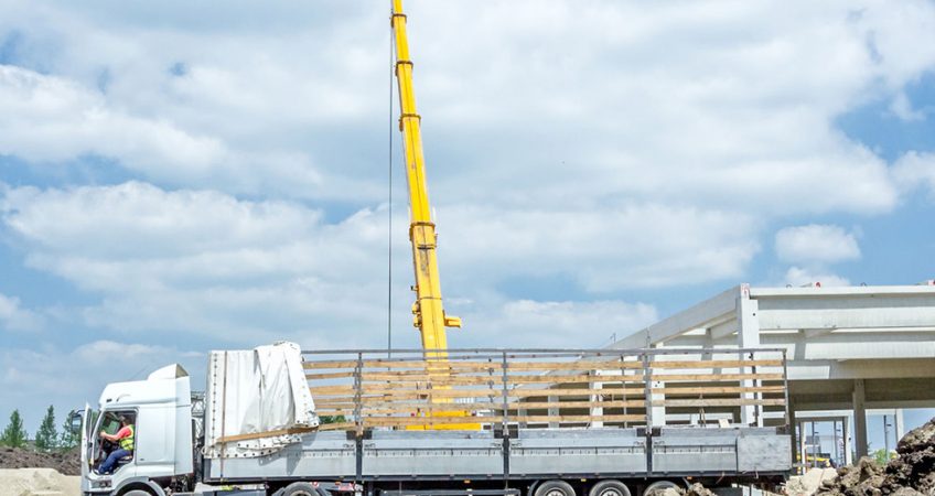Mobiele kraan tilt een stalen constructiebalk uit een vrachtwagen bij de bouw van een bedrijfshal